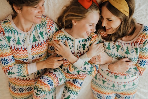 Three people are lying down on a white surface, all dressed in matching colorful patterned pajamas. They appear to be smiling and interacting warmly with each other.