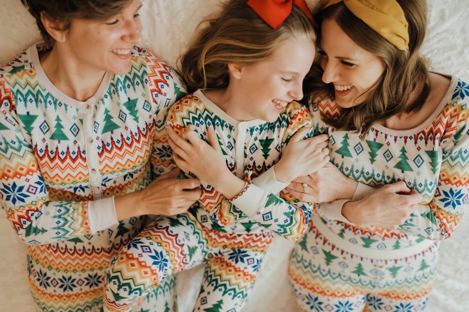 Three people are lying down on a white surface, all dressed in matching colorful patterned pajamas. They appear to be smiling and interacting warmly with each other.