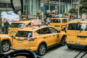 Multiple yellow taxis are clustered together in a busy urban environment. The scene includes a billboard displaying 'Bastille Day,' pedestrians in the background, and various street signs. The taxis are in various positions, some slightly angled, suggesting traffic congestion.