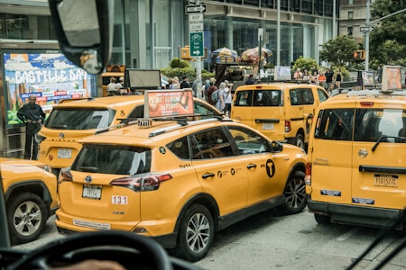 Multiple yellow taxis are clustered together in a busy urban environment. The scene includes a billboard displaying 'Bastille Day,' pedestrians in the background, and various street signs. The taxis are in various positions, some slightly angled, suggesting traffic congestion.