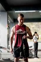 A man in a maroon tank top stands in a gym setting, holding what appears to be gym equipment. Behind him, a woman in a white t-shirt is seen in profile, standing with her hand near her mouth. Various gym equipment, including treadmills, is visible in the background.