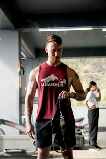 A man in a maroon tank top stands in a gym setting, holding what appears to be gym equipment. Behind him, a woman in a white t-shirt is seen in profile, standing with her hand near her mouth. Various gym equipment, including treadmills, is visible in the background.