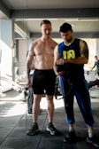 Two men are in a gym setting, where one is shirtless and wearing shorts, appearing to be in the process of a workout, while the other is dressed in athletic wear, holding a camera or similar device. They are standing on a gym mat with exercise equipment visible in the background.