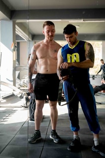 Two men are in a gym setting, where one is shirtless and wearing shorts, appearing to be in the process of a workout, while the other is dressed in athletic wear, holding a camera or similar device. They are standing on a gym mat with exercise equipment visible in the background.