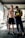 Two men are in a gym setting, where one is shirtless and wearing shorts, appearing to be in the process of a workout, while the other is dressed in athletic wear, holding a camera or similar device. They are standing on a gym mat with exercise equipment visible in the background.