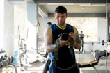 A man in a sleeveless shirt taking a photograph of himself in a gym mirror. The gym has various equipment such as exercise bikes and weights. Other people are visible exercising or on their phones in the background.