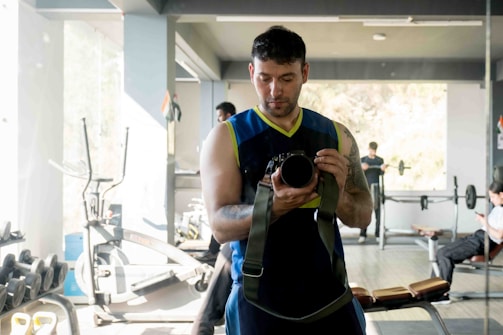 A man in a sleeveless shirt taking a photograph of himself in a gym mirror. The gym has various equipment such as exercise bikes and weights. Other people are visible exercising or on their phones in the background.