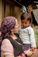 A woman holds a young child in her arms while standing outdoors. The woman wears a floral-patterned headscarf, traditional attire with gold embellishments, and several bracelets. The child looks down with a contemplative expression, wearing a light-colored shirt with designs. They are surrounded by a rural setting with laundry hanging in the background.