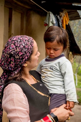 A woman holds a young child in her arms while standing outdoors. The woman wears a floral-patterned headscarf, traditional attire with gold embellishments, and several bracelets. The child looks down with a contemplative expression, wearing a light-colored shirt with designs. They are surrounded by a rural setting with laundry hanging in the background.