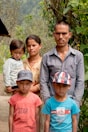 A group of five people, including two adults and three children, are standing outdoors. The adults, a man and a woman, are positioned in the back, with the woman holding a young child. Two older children, wearing caps, are standing in the front. The background is filled with greenery and a blurry view of trees and nature.