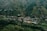 A small village nestled in a lush green valley surrounded by hills. The landscape is dotted with houses featuring colorful rooftops spread across the terrain, blending harmoniously with the natural environment. Dense patches of trees intersperse with cultivated fields, creating a patchwork of greenery.