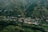 A small village nestled in a lush green valley surrounded by hills. The landscape is dotted with houses featuring colorful rooftops spread across the terrain, blending harmoniously with the natural environment. Dense patches of trees intersperse with cultivated fields, creating a patchwork of greenery.