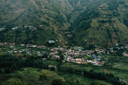 A small village nestled in a lush green valley surrounded by hills. The landscape is dotted with houses featuring colorful rooftops spread across the terrain, blending harmoniously with the natural environment. Dense patches of trees intersperse with cultivated fields, creating a patchwork of greenery.
