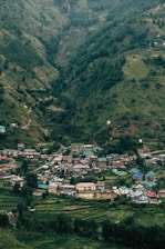 A small village nestled in a lush, green valley surrounded by hills. The village consists of colorful houses with some featuring distinct turquoise roofs. Dense forests cover the hillsides, indicating a rich natural environment.