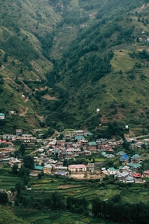 A small village nestled in a lush, green valley surrounded by hills. The village consists of colorful houses with some featuring distinct turquoise roofs. Dense forests cover the hillsides, indicating a rich natural environment.