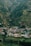 A small village nestled in a lush, green valley surrounded by hills. The village consists of colorful houses with some featuring distinct turquoise roofs. Dense forests cover the hillsides, indicating a rich natural environment.