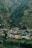 A small village nestled in a lush, green valley surrounded by hills. The village consists of colorful houses with some featuring distinct turquoise roofs. Dense forests cover the hillsides, indicating a rich natural environment.