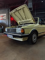 A vintage yellow convertible car with its hood open is parked in what appears to be a garage or workshop. The car has a classic design with rectangular headlights and a distinctive front grille, emblematic of older automotive styles. The environment is industrial, with brick walls, exposed piping, and signs, including one that reads 'CHAMPION'. The lighting is dim, with a red light casting a warm glow over part of the scene.