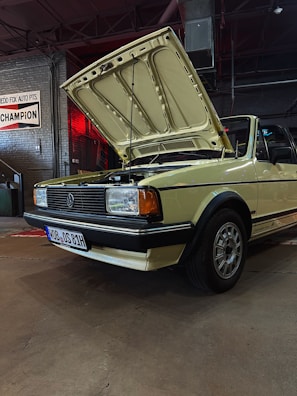 A vintage yellow convertible car with its hood open is parked in what appears to be a garage or workshop. The car has a classic design with rectangular headlights and a distinctive front grille, emblematic of older automotive styles. The environment is industrial, with brick walls, exposed piping, and signs, including one that reads 'CHAMPION'. The lighting is dim, with a red light casting a warm glow over part of the scene.