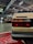 The image depicts an indoor car exhibition focusing on the rear of a beige Volkswagen vehicle prominently in the foreground. In the background, other vehicles, including a race car, are visible amidst the subdued lighting and various automotive signs. The setting gives the impression of a classic car show or a garage.