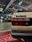 The image depicts an indoor car exhibition focusing on the rear of a beige Volkswagen vehicle prominently in the foreground. In the background, other vehicles, including a race car, are visible amidst the subdued lighting and various automotive signs. The setting gives the impression of a classic car show or a garage.