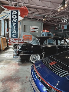 An indoor setting of a vintage garage featuring classic cars and retro signs. Prominent is a large red and blue Pontiac sign with other advertisements like Avon Tyres and Champion on the wall. Displayed are a black vintage car with whitewall tires and a modern blue Porsche, contributing to an interesting blend of old and new automotive aesthetics.