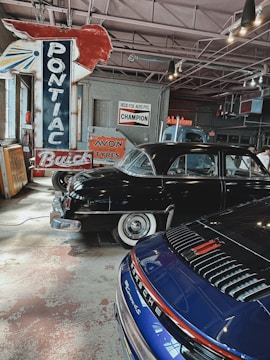 An indoor setting of a vintage garage featuring classic cars and retro signs. Prominent is a large red and blue Pontiac sign with other advertisements like Avon Tyres and Champion on the wall. Displayed are a black vintage car with whitewall tires and a modern blue Porsche, contributing to an interesting blend of old and new automotive aesthetics.
