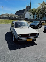 A vintage Volkswagen car is parked in a lot with a dark gray asphalt surface. In the background, a stone church with Gothic architecture is visible, surrounded by a wooden fence and adjacent to residential houses. The car has a yellow license plate with the number 3190 and is well-lit under clear blue skies, casting distinct shadows.