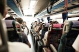 Passengers are seated inside a bus, facing forward. The view shows multiple rows of seats occupied by people with varying hairstyles and clothing. The bus interior is well-lit, with overhead storage and windows on both sides.
