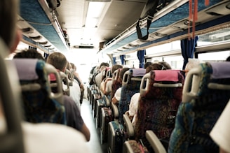 Passengers are seated inside a bus, facing forward. The view shows multiple rows of seats occupied by people with varying hairstyles and clothing. The bus interior is well-lit, with overhead storage and windows on both sides.
