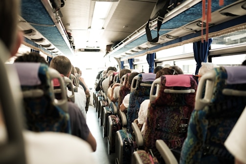 Passengers are seated inside a bus, facing forward. The view shows multiple rows of seats occupied by people with varying hairstyles and clothing. The bus interior is well-lit, with overhead storage and windows on both sides.