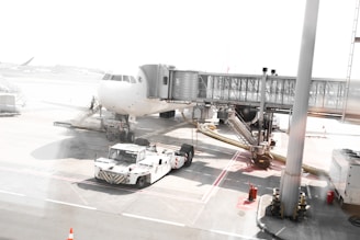 A commercial airplane is parked at an airport gate with a jet bridge connected to it for passenger boarding. Ground support vehicles and equipment are visible around the aircraft, including a pushback tug. The scene is brightly lit with sunlight, highlighting the white and grey tones of the airport tarmac and infrastructure.