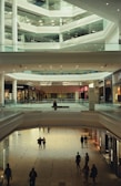 A spacious, multi-level interior of a modern shopping mall with people walking on a tiled floor. The architecture is characterized by sleek lines and glass railings. Stores line the sides of the mall, and several shoppers are visible at different levels and in the foreground.