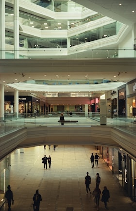 A spacious, multi-level interior of a modern shopping mall with people walking on a tiled floor. The architecture is characterized by sleek lines and glass railings. Stores line the sides of the mall, and several shoppers are visible at different levels and in the foreground.