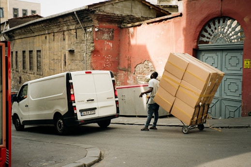 A person is maneuvering a hand truck loaded with large boxes on a street. The individual is dressed casually and appears to be delivering or transporting the parcels. A white van is parked nearby, partially blocking the road. The surroundings include an old, weathered building with a red, peeling wall and a decorative blue door.