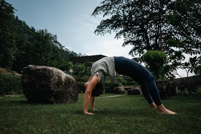 A person is performing a backbend yoga pose on a grassy field, surrounded by greenery and large rocks. In the background, there are trees and a partially cloudy sky.