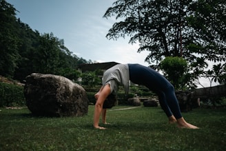 A person is performing a backbend yoga pose on a grassy field, surrounded by greenery and large rocks. In the background, there are trees and a partially cloudy sky.