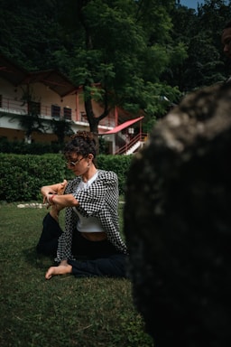 A person practicing yoga or stretching on a green lawn in a lush, wooded area with a building featuring red accents in the background.