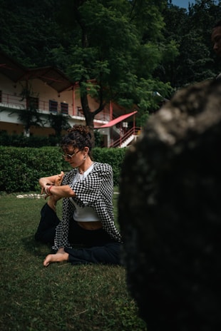 A person practicing yoga or stretching on a green lawn in a lush, wooded area with a building featuring red accents in the background.