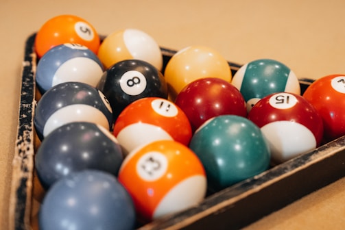 A set of pool balls arranged in a triangular rack on a billiard table. The balls display a variety of colors, including orange, red, blue, black, and white, each with numbers printed on them.