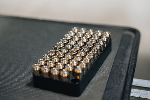 A black tray holds a neatly arranged grid of shiny bullet casings, each reflecting light. The arrangement is orderly, suggesting a sense of preparation or organization.