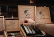 A hand reaches into a box of cigars with artistic branding on a shelf. The box features a prominent profile of a man's silhouette alongside the logo and text, indicating the brand's focus on craftsmanship. The wooden display boxes are intricately designed, showcasing premium cigars.