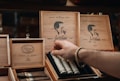 A hand reaches into a box of cigars with artistic branding on a shelf. The box features a prominent profile of a man's silhouette alongside the logo and text, indicating the brand's focus on craftsmanship. The wooden display boxes are intricately designed, showcasing premium cigars.