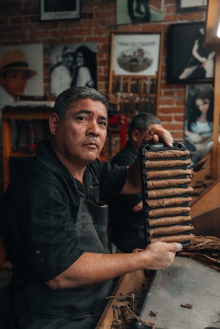 A man in a workshop setting holds up a tray filled with rolled cigars. The background features several framed black-and-white photographs on a brick wall. The man wears a dark apron and appears focused on his task.