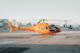 A bright orange helicopter is parked on a tarmac at an airfield, surrounded by a clear sky and distant mountains. In the background, there are modern buildings and a jet plane near the fence.