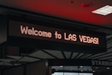 A digital sign displays the text 'Welcome to LAS VEGAS!' in bright lights, positioned indoors with a visible ceiling featuring geometric patterns and some lighting fixtures.