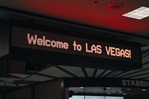 A digital sign displays the text 'Welcome to LAS VEGAS!' in bright lights, positioned indoors with a visible ceiling featuring geometric patterns and some lighting fixtures.