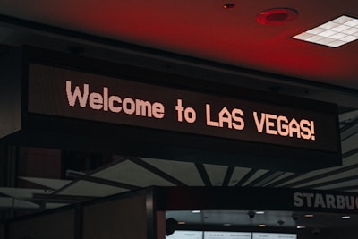 A digital sign displays the text 'Welcome to LAS VEGAS!' in bright lights, positioned indoors with a visible ceiling featuring geometric patterns and some lighting fixtures.