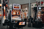 A collection of slot machines is situated inside a building with bright, colorful screens displaying various games and gaming themes. A person is seated nearby, seemingly relaxed with luggage beside them, and the atmosphere is lively with electronic displays.