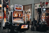 A collection of slot machines is situated inside a building with bright, colorful screens displaying various games and gaming themes. A person is seated nearby, seemingly relaxed with luggage beside them, and the atmosphere is lively with electronic displays.